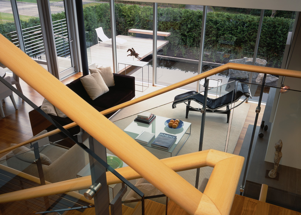 Living room in a Vancouver custom home features large windows, a black chaise lounge, sofa, and glass coffee table near the staircase—showcasing quality design and style.