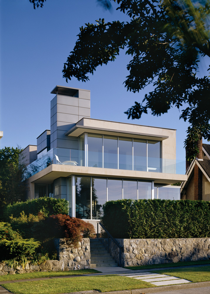 Two-story house with large glass windows, stone wall, and balcony, surrounded by greenery. Built by a leading general contractor in British Columbia.