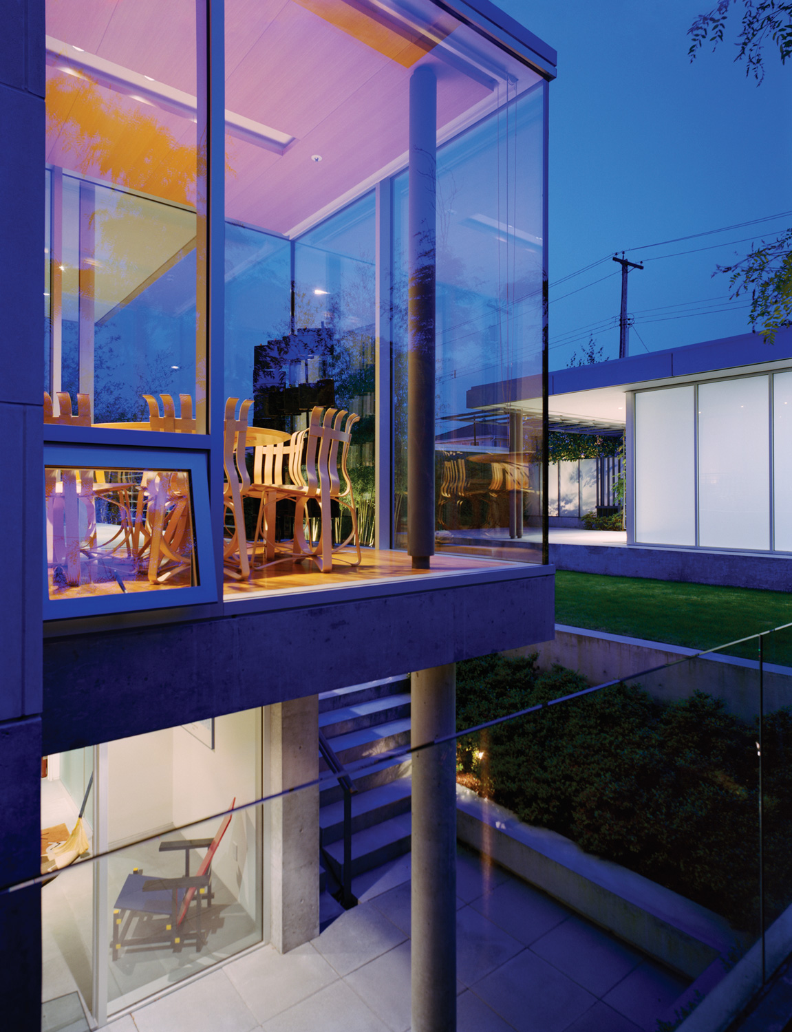 Glass house at dusk with stacked rooms showcases striking architecture, visible chairs, and outdoor greenery—an inspiring example of quality custom homes in Vancouver