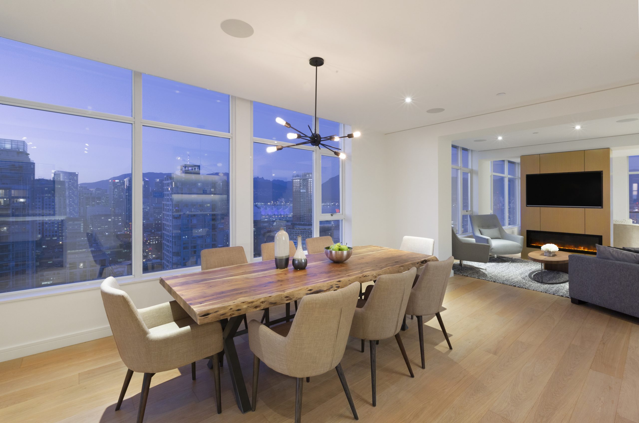 Elegant dining room in a custom home featuring a wooden table, beige chairs, large windows, and a stunning city view at dusk—showcasing refined architecture in British Columbia.