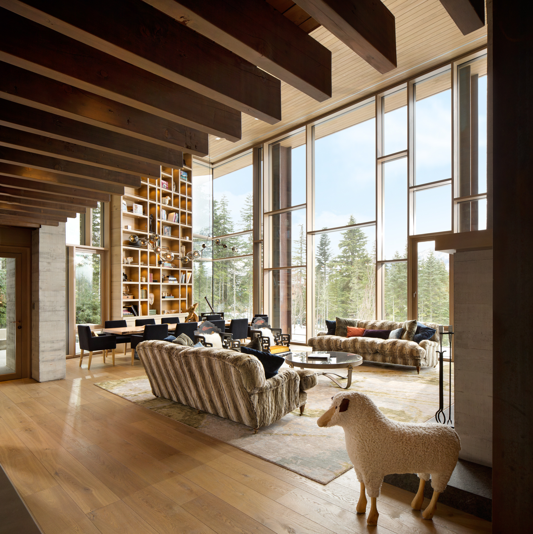 Living room with large windows, wooden beams, cozy sofas, tall bookshelf, and a decorative sheep statue—meticulously crafted by a leading General Contractor in BC.