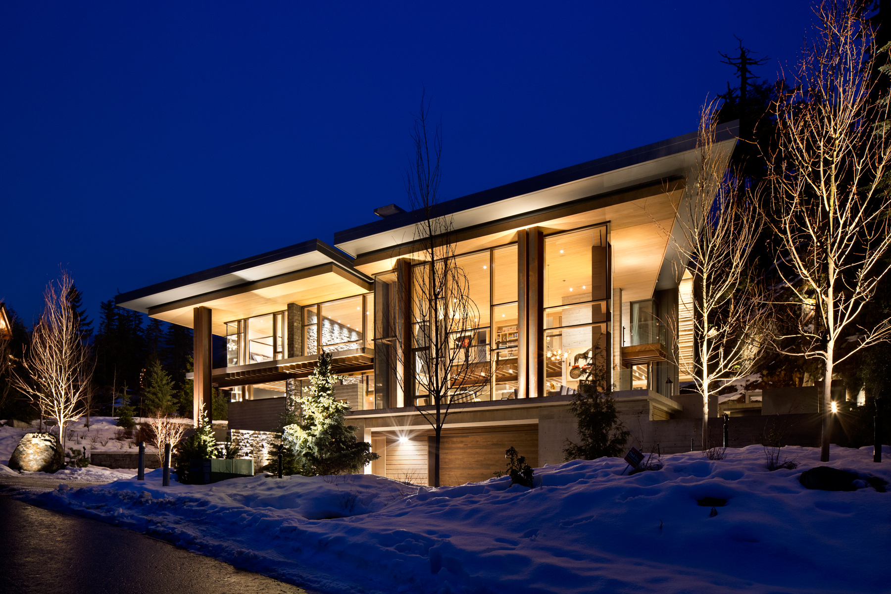 House with large windows, warmly lit, surrounded by snow and trees at night. Expertly built by a leading BC general contractor, this construction blends contemporary design with the natural landscape for breathtaking results.