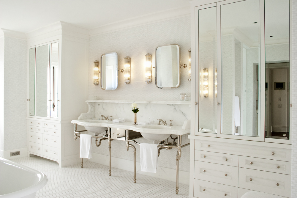 Elegant white bathroom with double sinks, large mirrors, glass cabinets, and bright vanity lights—showcasing the quality craftsmanship found in Vancouver custom homes.