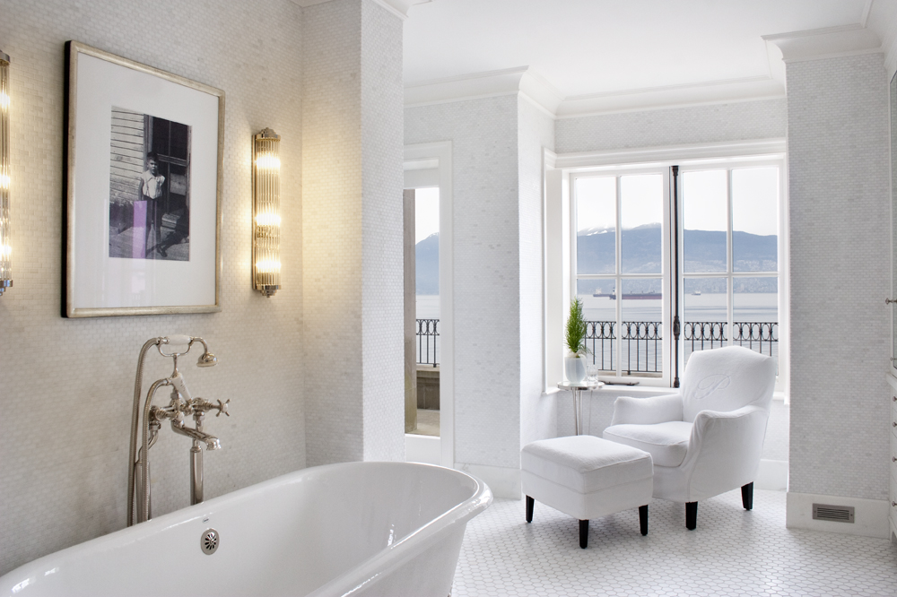Bright, elegant bathroom with a white armchair, bathtub, wall art, and large window overlooking the mountains of Vancouver, British Columbia, exuding quality and style.