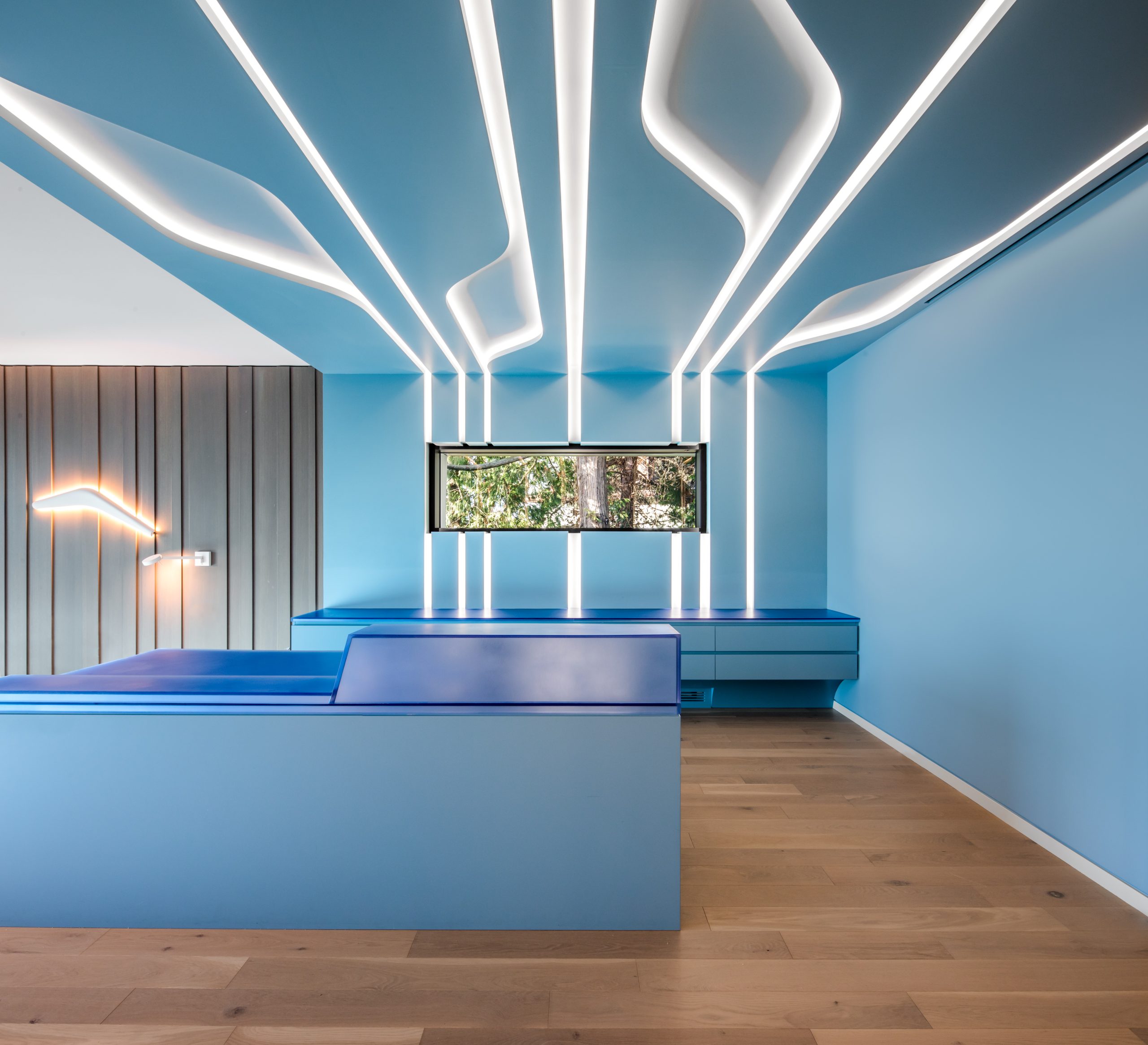 Futuristic blue room by a leading construction company in British Columbia, featuring wooden floor, white LED light strips on ceiling and wall, and a window with views of trees.