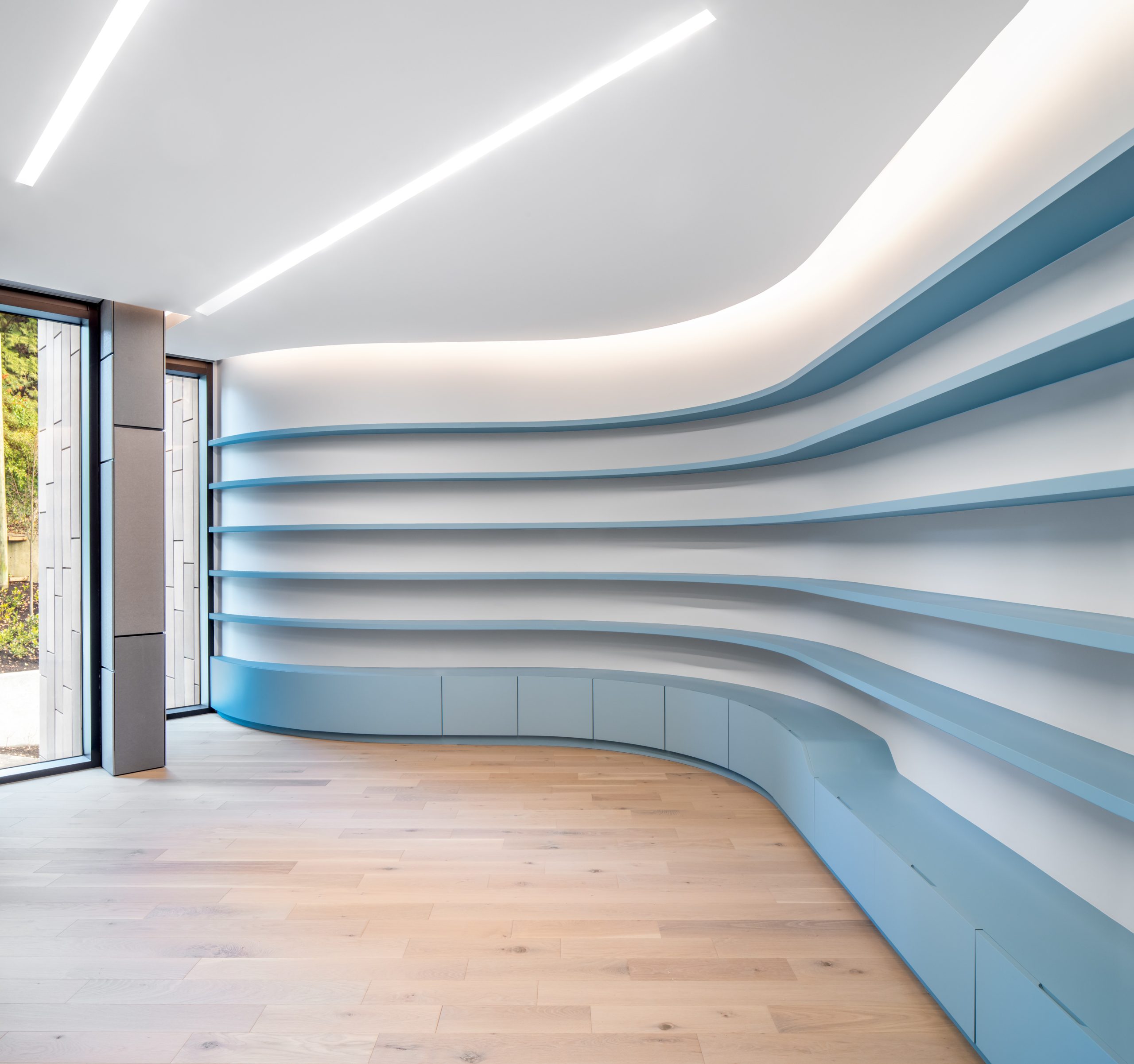 Room by a British Columbia general contractor, featuring curved, empty blue shelves, wood floor, and large windows letting in natural light.