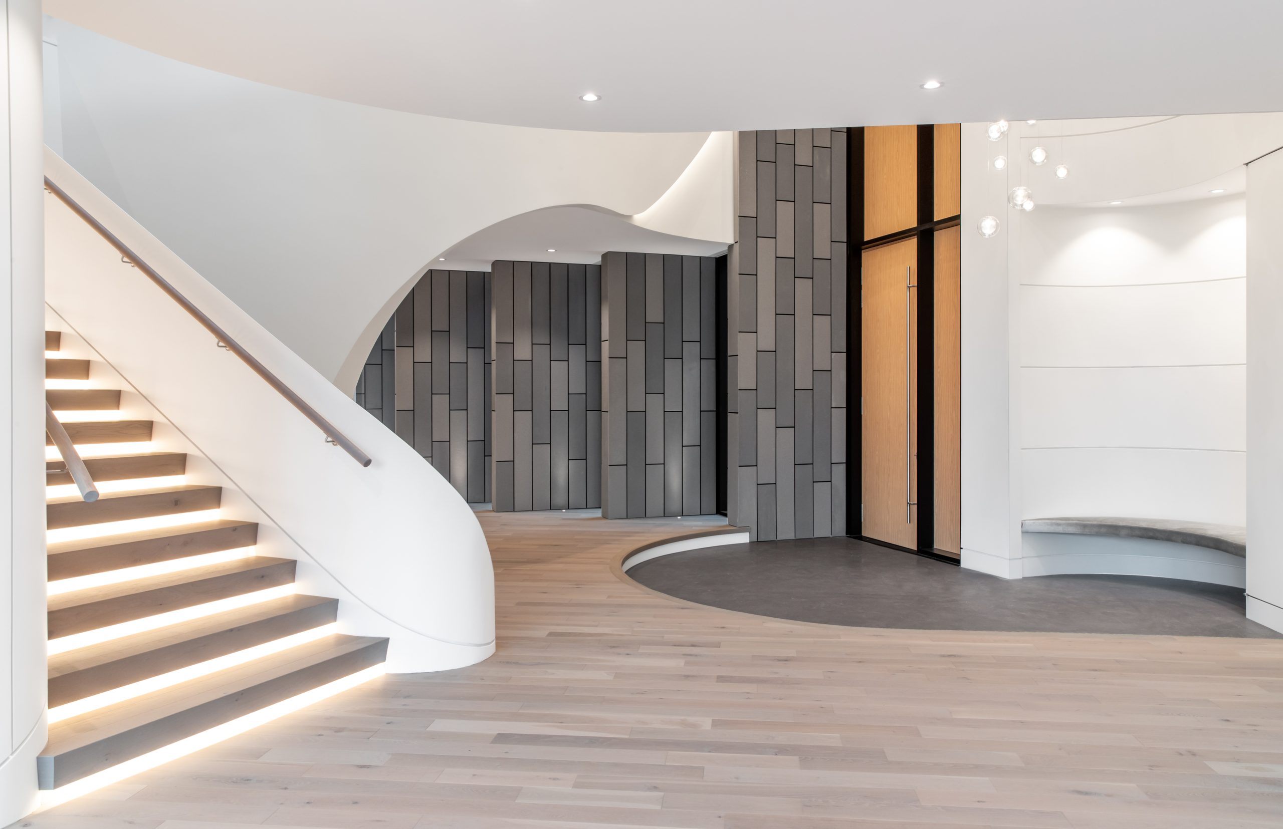 Home lobby by a reputable company, featuring a curved staircase, wood flooring, grey panel walls, and hanging lights—an elegant welcome to custom homes in British Columbia.