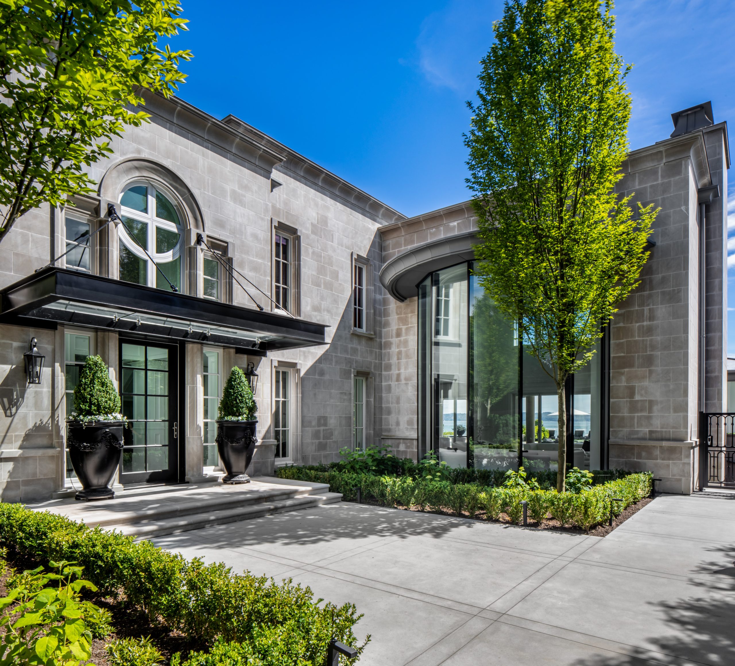 Large modern stone house with tall windows, manicured shrubs, and a bright blue sky showcases stunning architecture often seen in British Columbia.