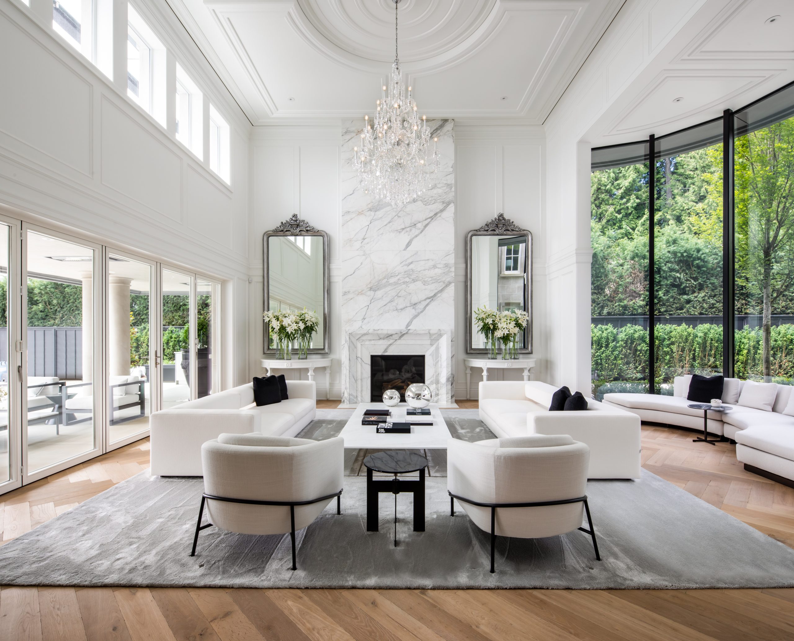 Elegant, spacious living room with quality white sofas, marble fireplace, chandeliers, and large windows showcasing the beautiful greenery of British Columbia’s landscape.
