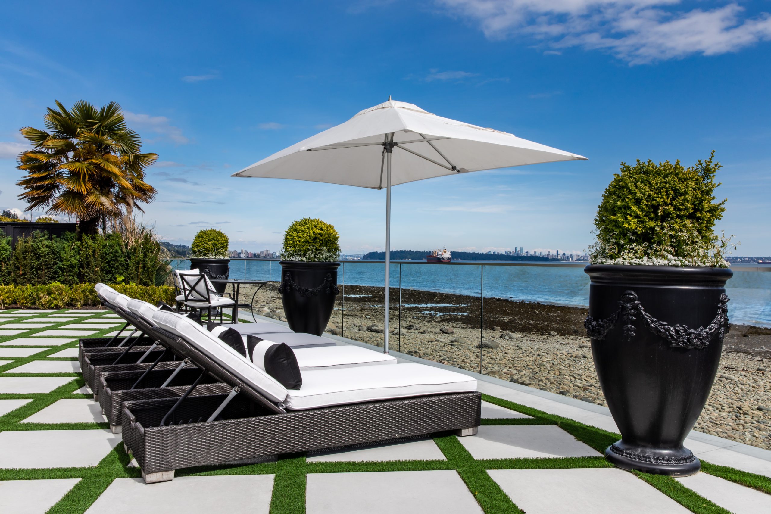 Lounge chairs and an umbrella on a patio by the water, surrounded by large potted plants and a clear sky—an inviting outdoor space inspired by the stunning architecture of custom homes in British Columbia.