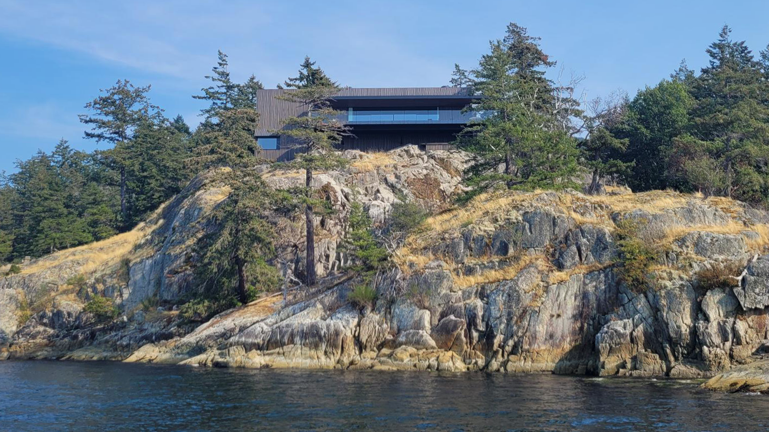 House, built by a leading construction company, sits atop a rocky cliff surrounded by trees and overlooks a body of water under a clear sky.