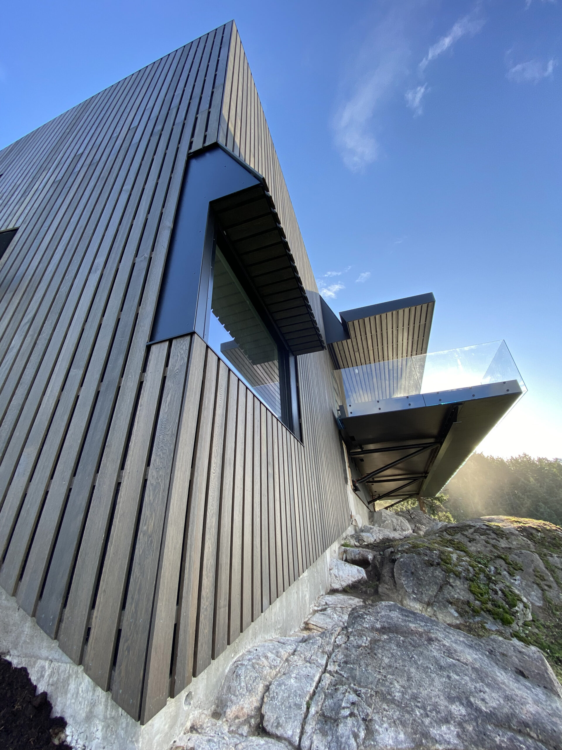 Quality modern house with vertical wooden siding, large windows, and glass balcony, built on a rocky slope in Vancouver, British Columbia.