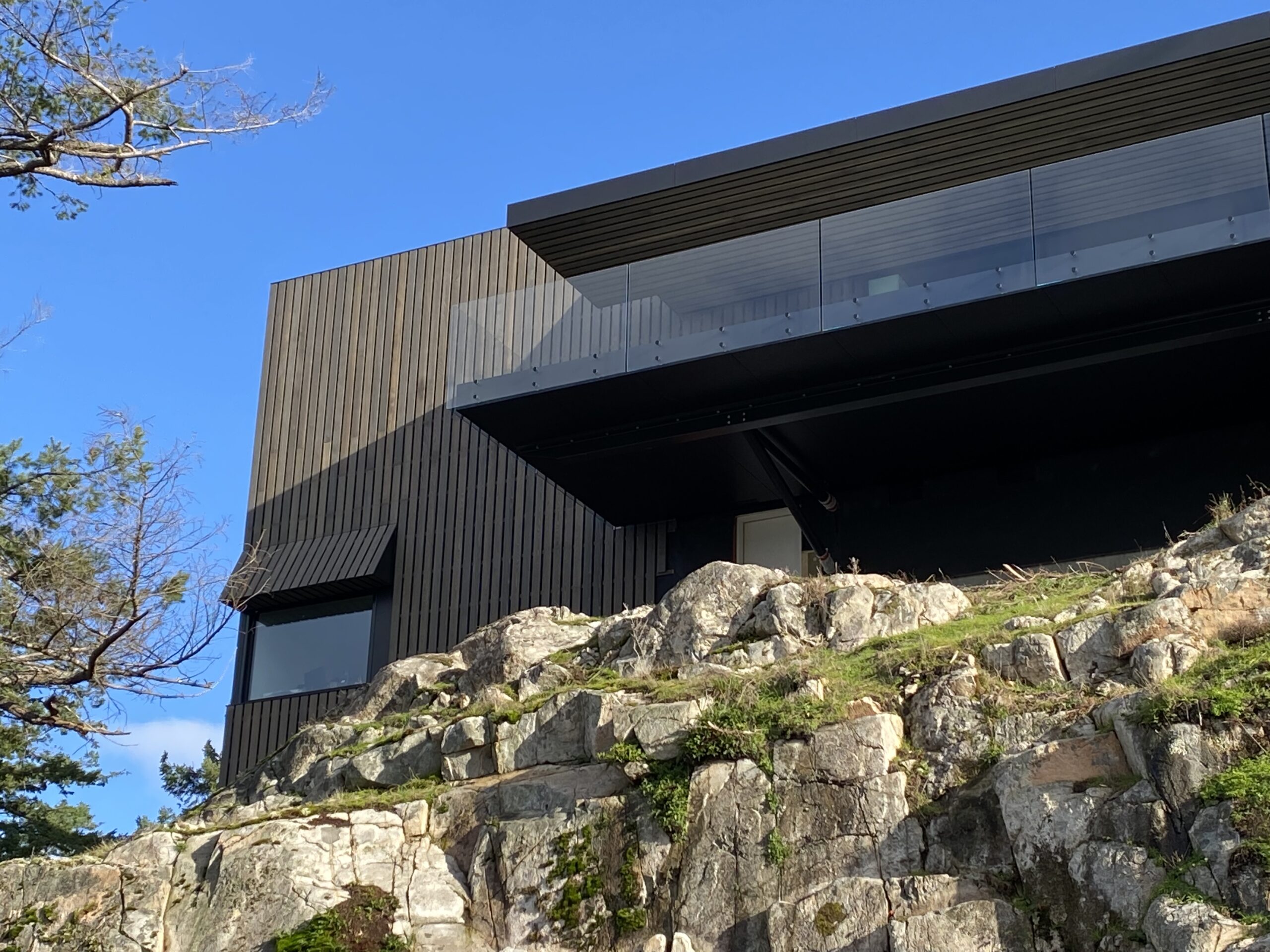 Modern dark wood house with large glass balcony, built on rocky terrain under a clear Vancouver sky by a reputable construction team specializing in custom homes.