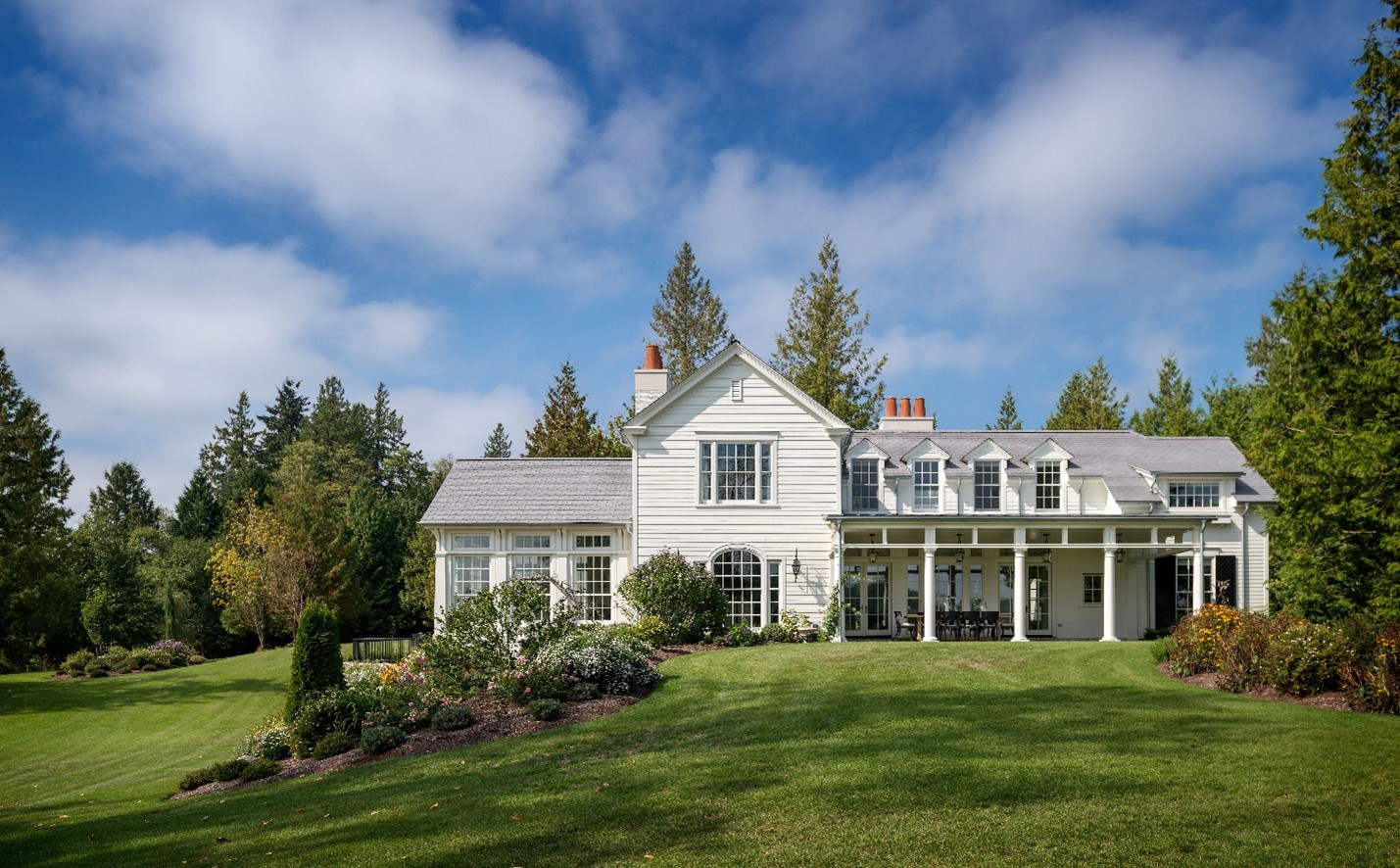 White two-story house with a large green lawn, garden, and trees under a partly cloudy sky.