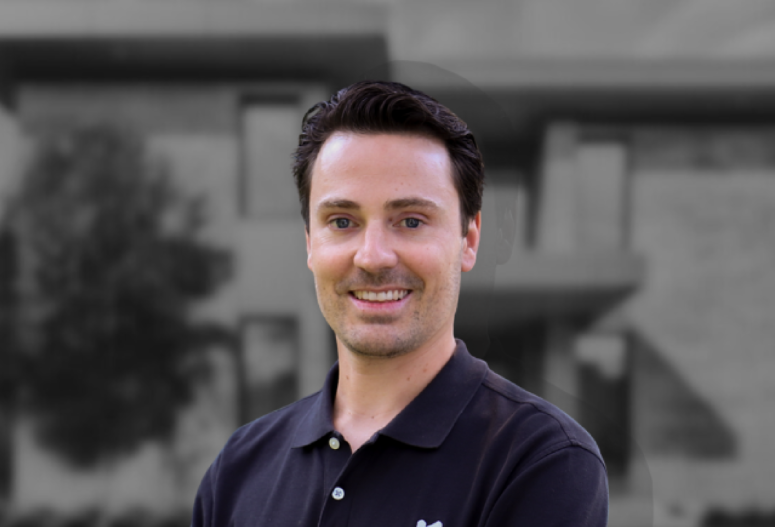 Smiling man with dark hair in a black polo shirt stands in front of a blurred building background.